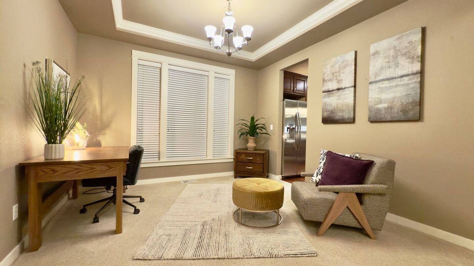 A well-lit room with a wooden desk, a comfortable armchair, a round ottoman, and a large window with blinds.