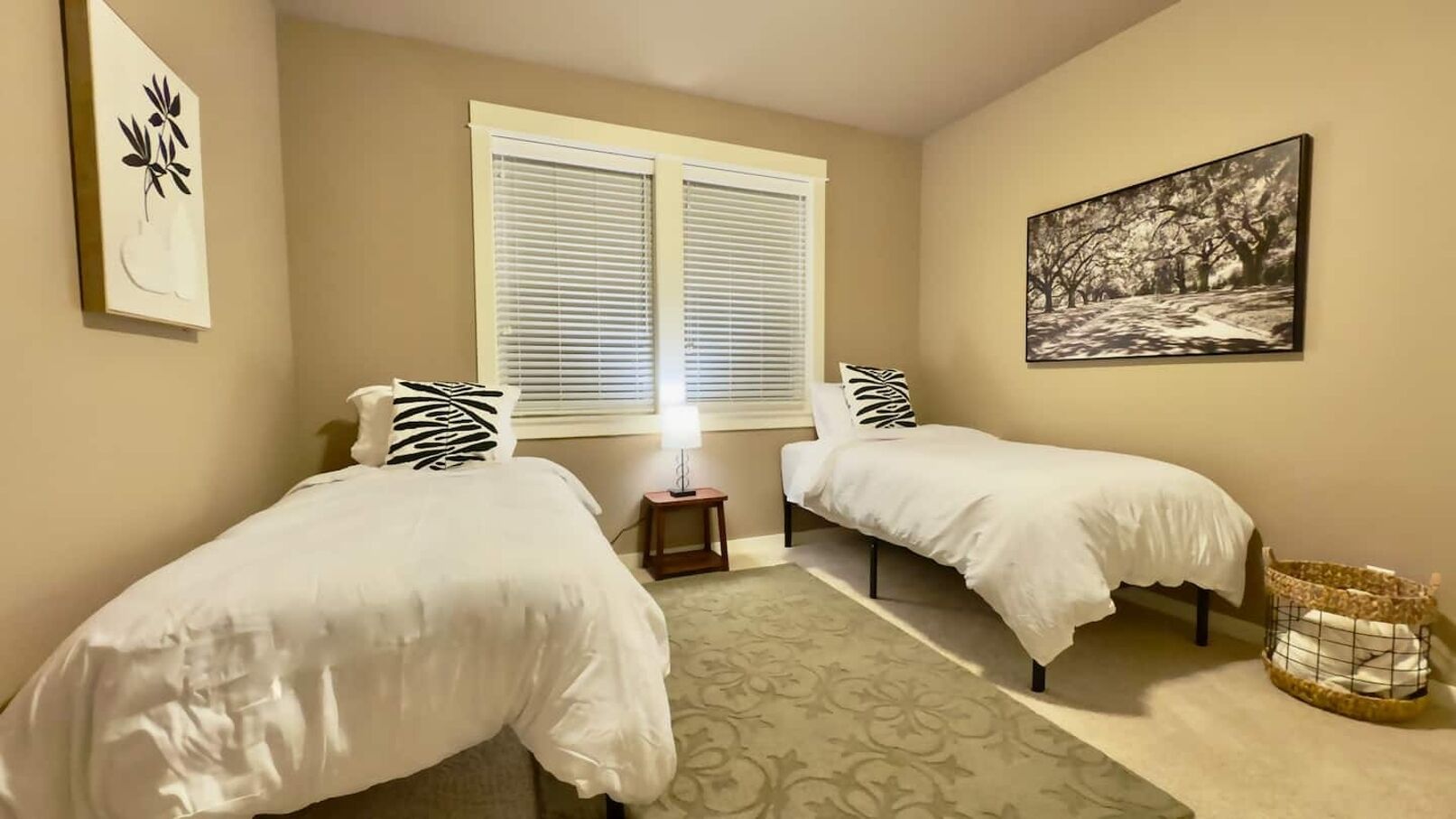 Een slaapkamer met twee éénpersoonsbedden, een raam met zonwering, een nachtkastje met een lamp, een lijst met een foto aan de muur en een klein mandje op de vloer.