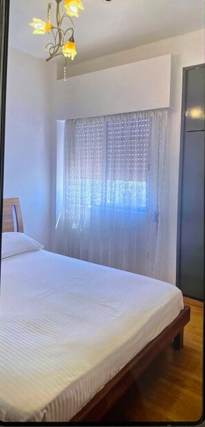 Ein Schlafzimmer mit Bett, einem Holzkopfteil, einer Chandelierleuchte und einem Fenster mit durchsichtigen Vorhängen.