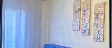Een woonkamer met een blauwe bank, een ronde salontafel en een kroonluchter. Daarboven hangen drie kunstwerken.