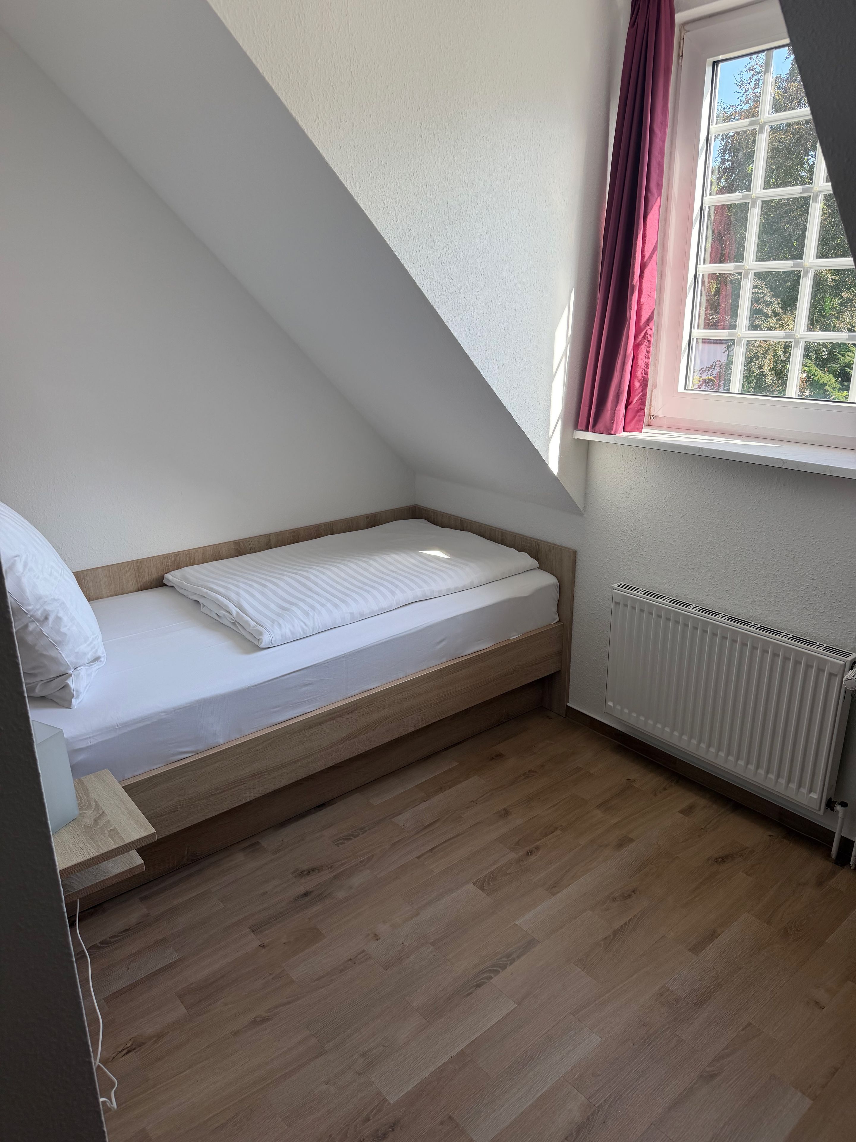 A small, sloped ceiling room with a single bed, a window with curtains, and a radiator.