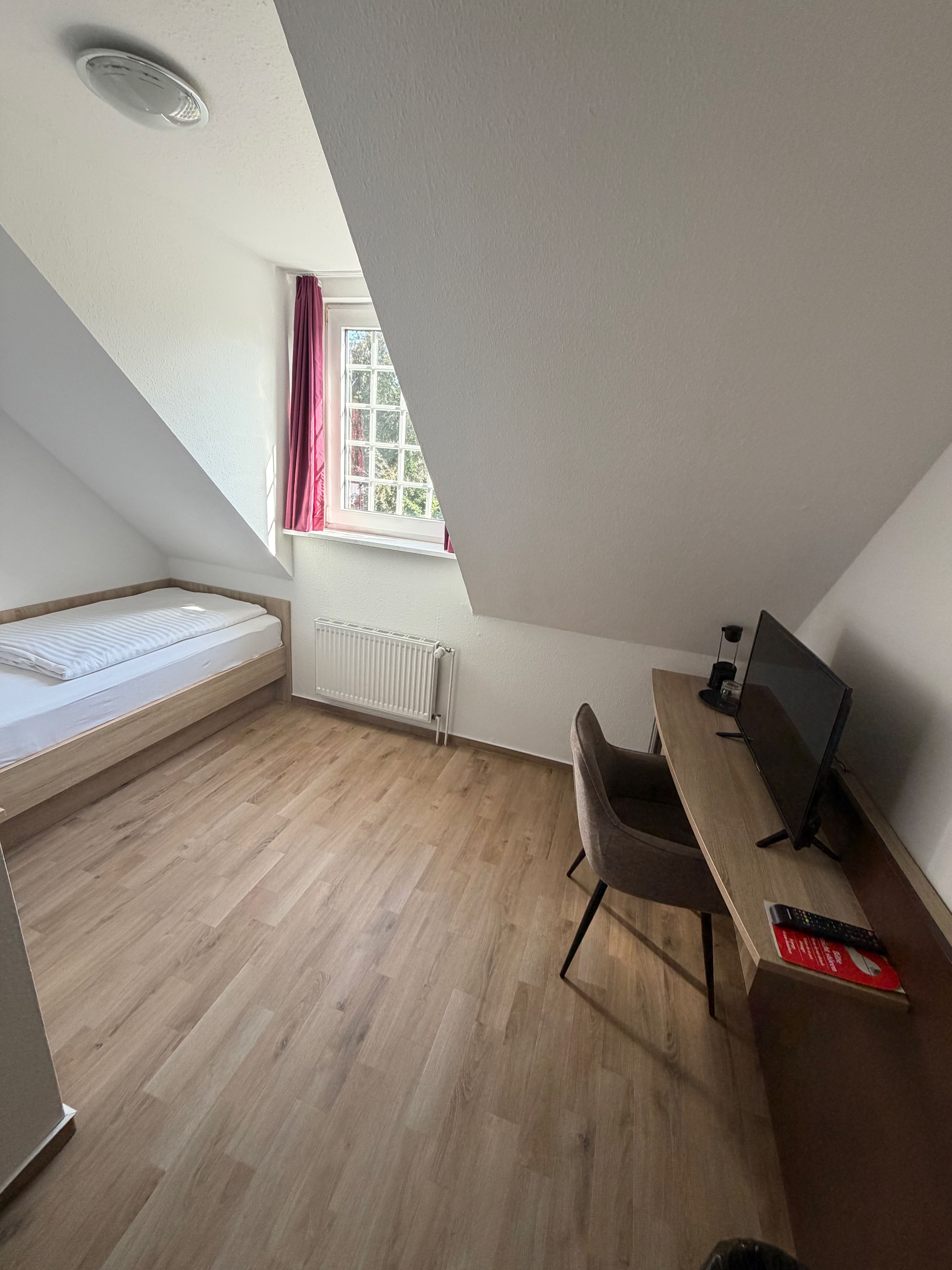 A room with a sloped ceiling, a bed, a desk with a computer, and a window with curtains.