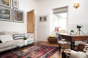 Una sala de estar con una mesa de centro de vidrio, un tapete estampado, un sofá con almohadones a rayas, una silla de mimbre y una mesa de comedor con patas de trípode.
