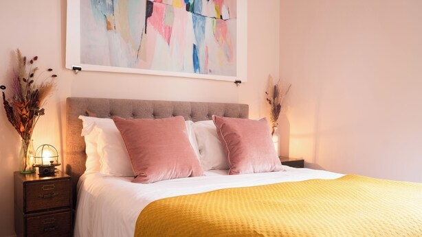 Ein ordentlich bezogenes Bett mit einer gelben Decke, einem grau gepolsterten Kopfteil und rosafarbenen Kissen. Ein Nachttisch mit Lampe und einer Vase mit getrockneten Blumen steht daneben. Über dem Bett hängt ein farbenfrohes abstraktes Gemälde.