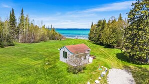 A small stone house with a red roof, surrounded by a well-maintained lawn and trees, near a body of water. - Million Dollar View | Lakefront Studio Cottage w/ Mackinac Bridge & Island Views (Cheboygan)