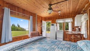 A cabin with a wooden interior, a bed with a blue and white quilt, a kitchenette with a stove and refrigerator, and a view of the outdoors through a window. - Million Dollar View | Lakefront Studio Cottage w/ Mackinac Bridge & Island Views (Cheboygan)
