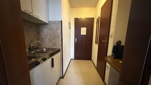 A narrow hallway with a kitchen area on the left, featuring a sink and cabinets. A door with a sign is visible in the distance.