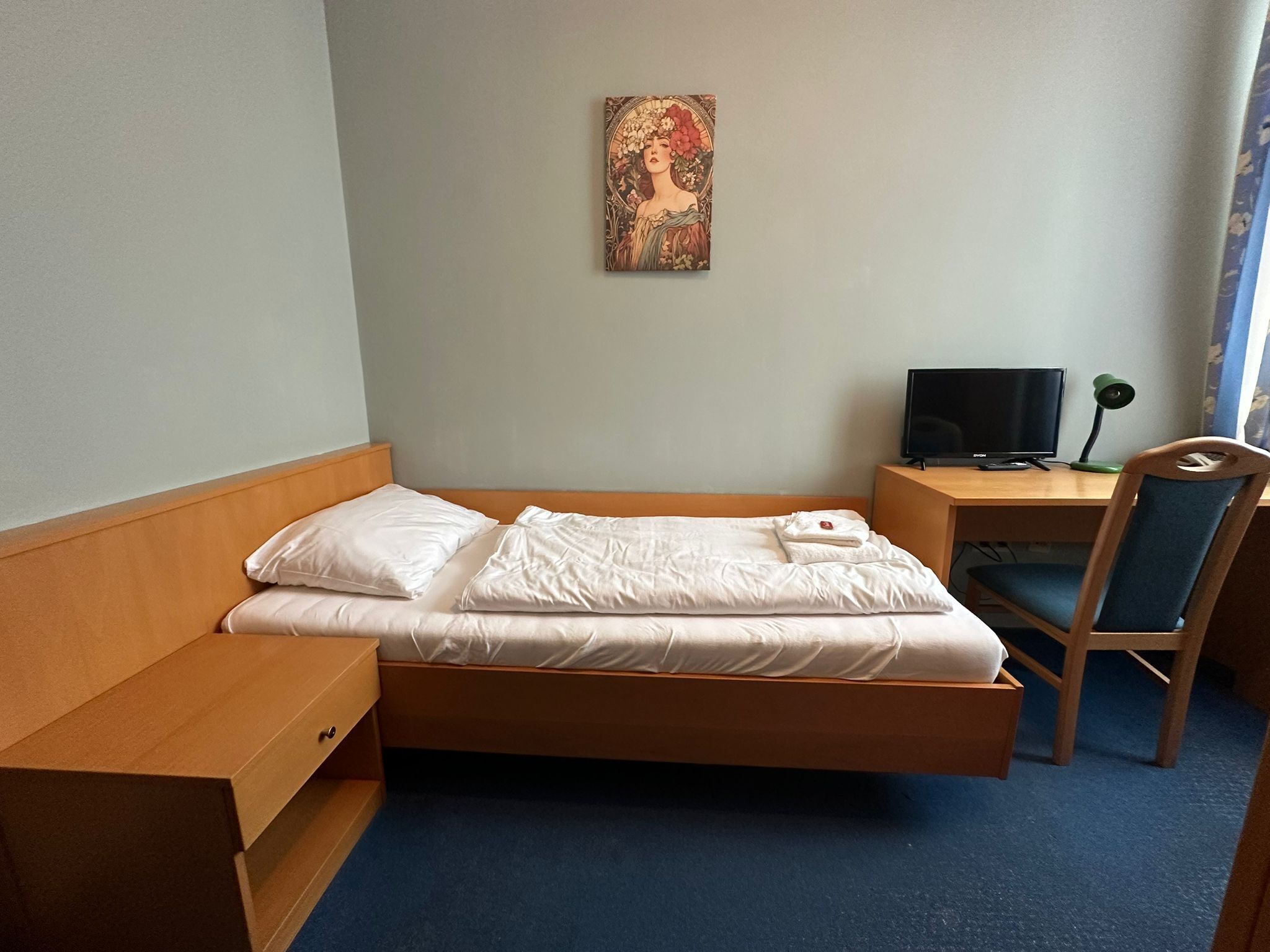 A single bed with white linens, a wooden headboard, a bedside table with a lamp, a desk with a computer, and a chair.