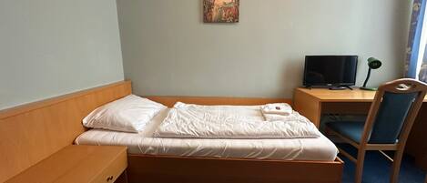 A single bed with white linens, a wooden headboard, a bedside table with a lamp, a desk with a computer, and a chair.