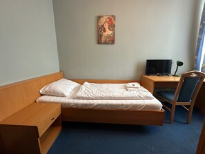 Ein Einzelbett mit weißer Bettwäsche, ein Holz-Kopfteil, ein Nachttisch mit Lampe, ein Schreibtisch mit Computer und ein Stuhl.