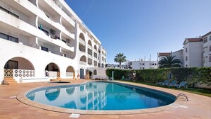 A swimming pool area with a white building in the background, a palm tree, and a clear sky. - Spacious apartment in the heart of Albufeira (Albufeira)