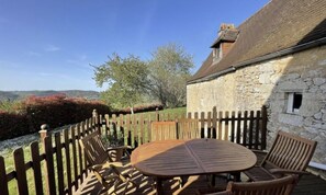 Un bâtiment en pierre doté d’une terrasse en bois, d’une table ronde et de deux chaises.