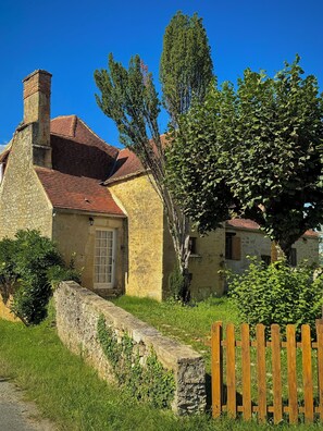 Une maison en pierre avec un toit recouvert de tuiles rouges, une clôture en bois et un grand arbre au premier plan.