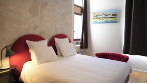 Hotellihuone, jossa on suuri sänky, punainen pääty, valkoiset vuodevaatteet, yöpöytä lampun kanssa ja seinällä kuvaus rannikkokaupungista.