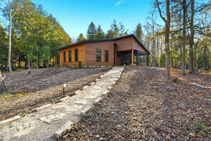 A modern wooden cabin with a stone pathway leading to the entrance, surrounded by a forest.