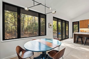 Un espace repas moderne avec une table ronde, des chaises et de grandes fenêtres donnant sur une forêt.