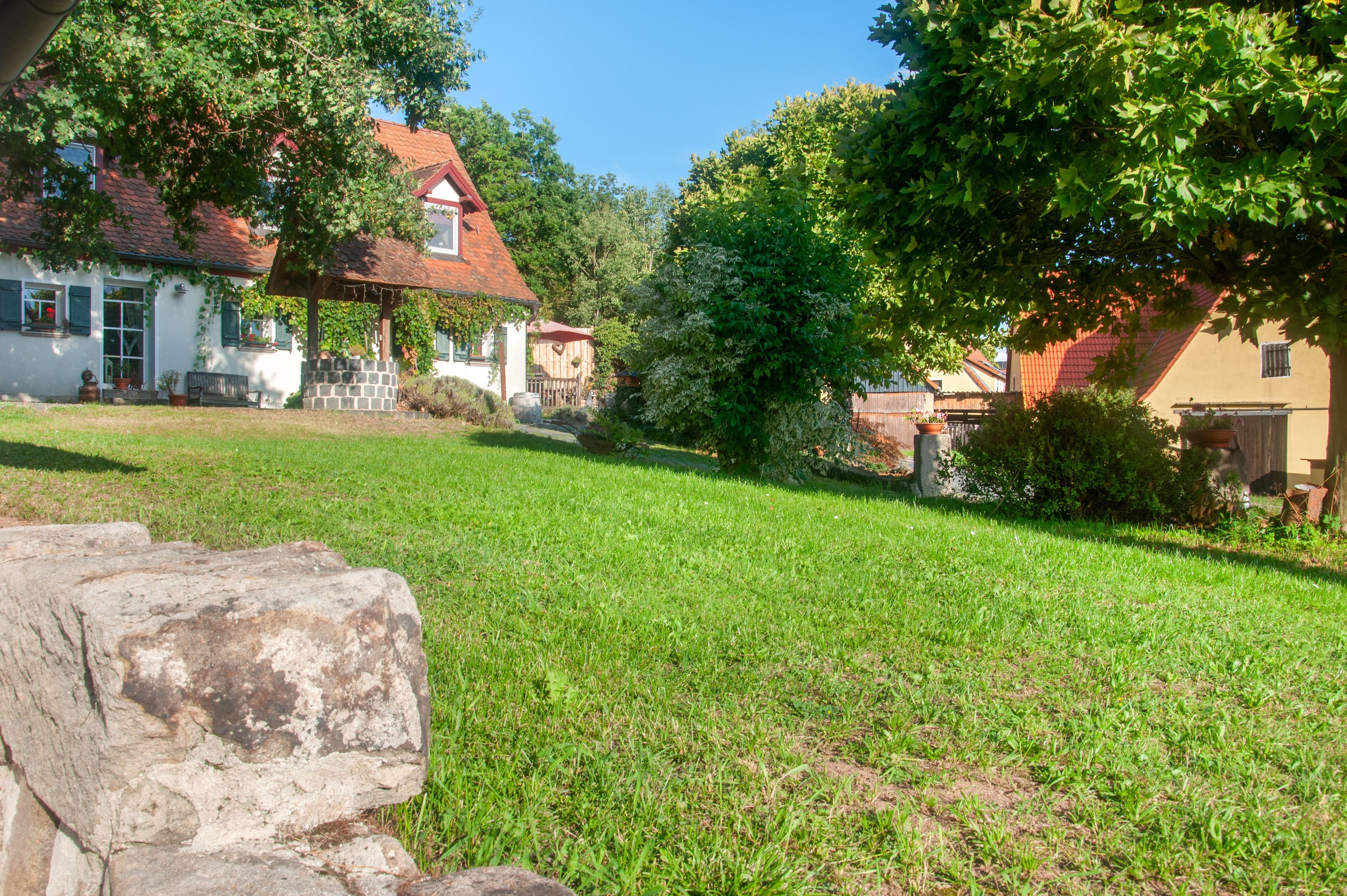 A grassy yard with a stone wall, a white building with a red roof, and a tree.