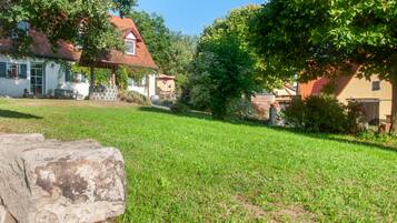 A grassy yard with a stone wall, a white building with a red roof, and a tree.