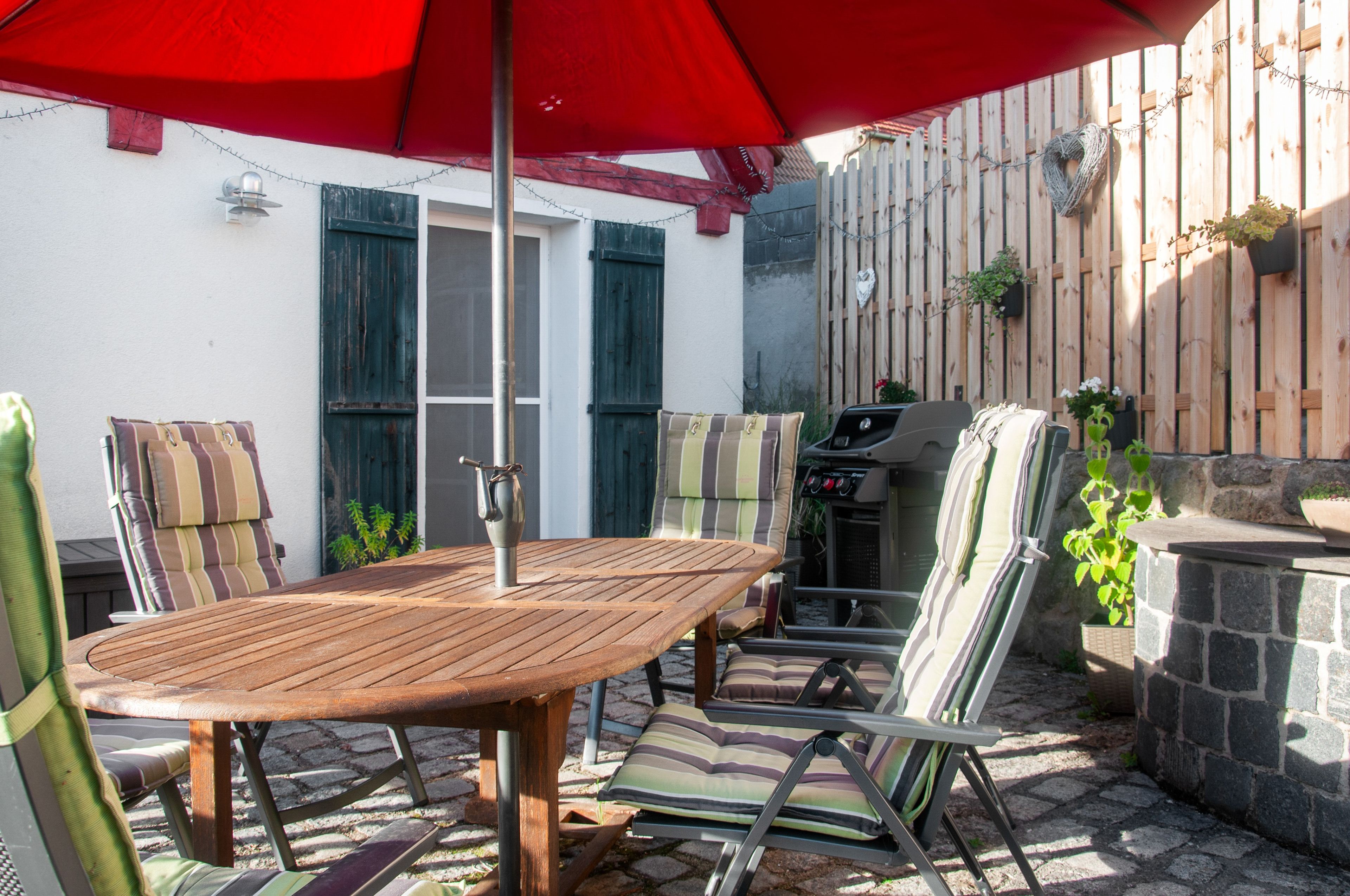 An outdoor patio with a wooden table and chairs, a barbecue grill, and a red umbrella.