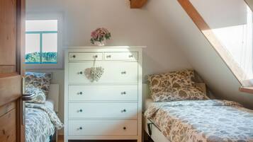 Une chambre avec une commode blanche, deux lits avec une literie à motifs, une fenêtre donnant sur des arbres et une poutre en bois au plafond.
