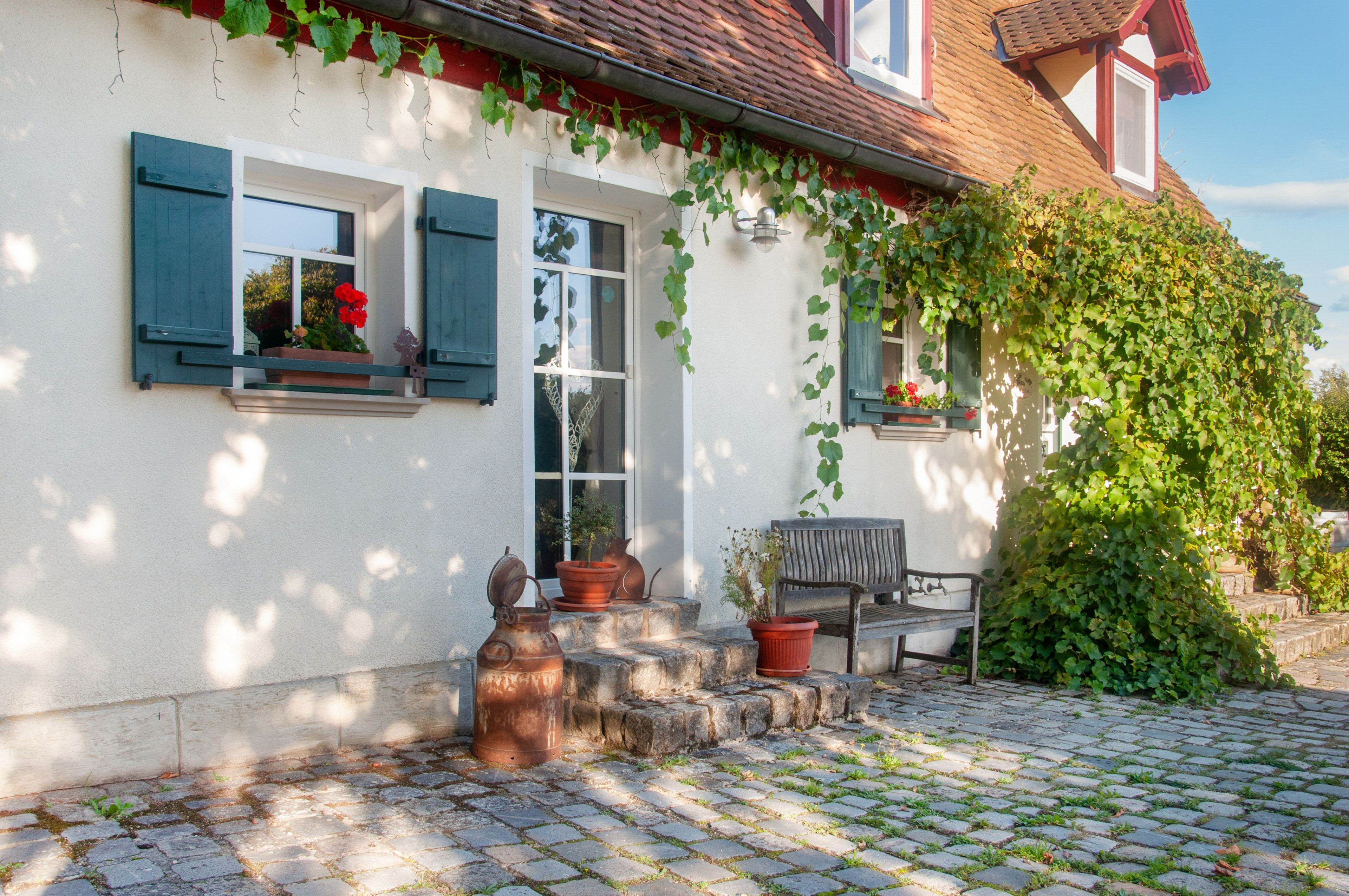 A house with a white exterior, green shutters, and a brick roof. There is a bench and potted plants on the front porch.