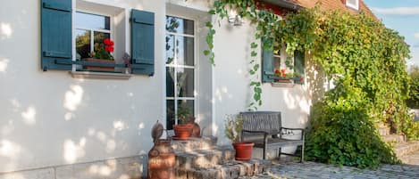 Una casa con facciata bianca, persiane verdi e tetto in mattoni. Il portico presenta una panchina e piante in vaso.