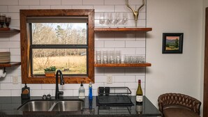 A kitchen with a large window, stainless steel sink, wooden shelves, and a wine bottle. - Grace Farmhouse-12 ac,  hot tub, fire pit (Menlo)