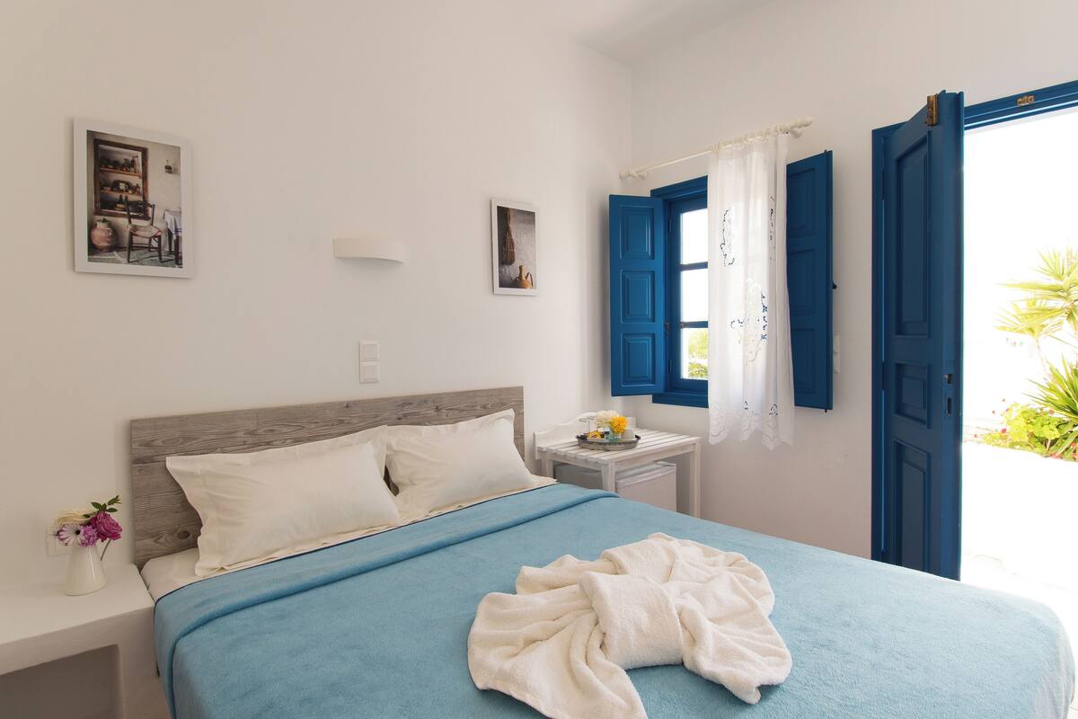 A bedroom with a bed, a nightstand, a small table, and a window with blue shutters.