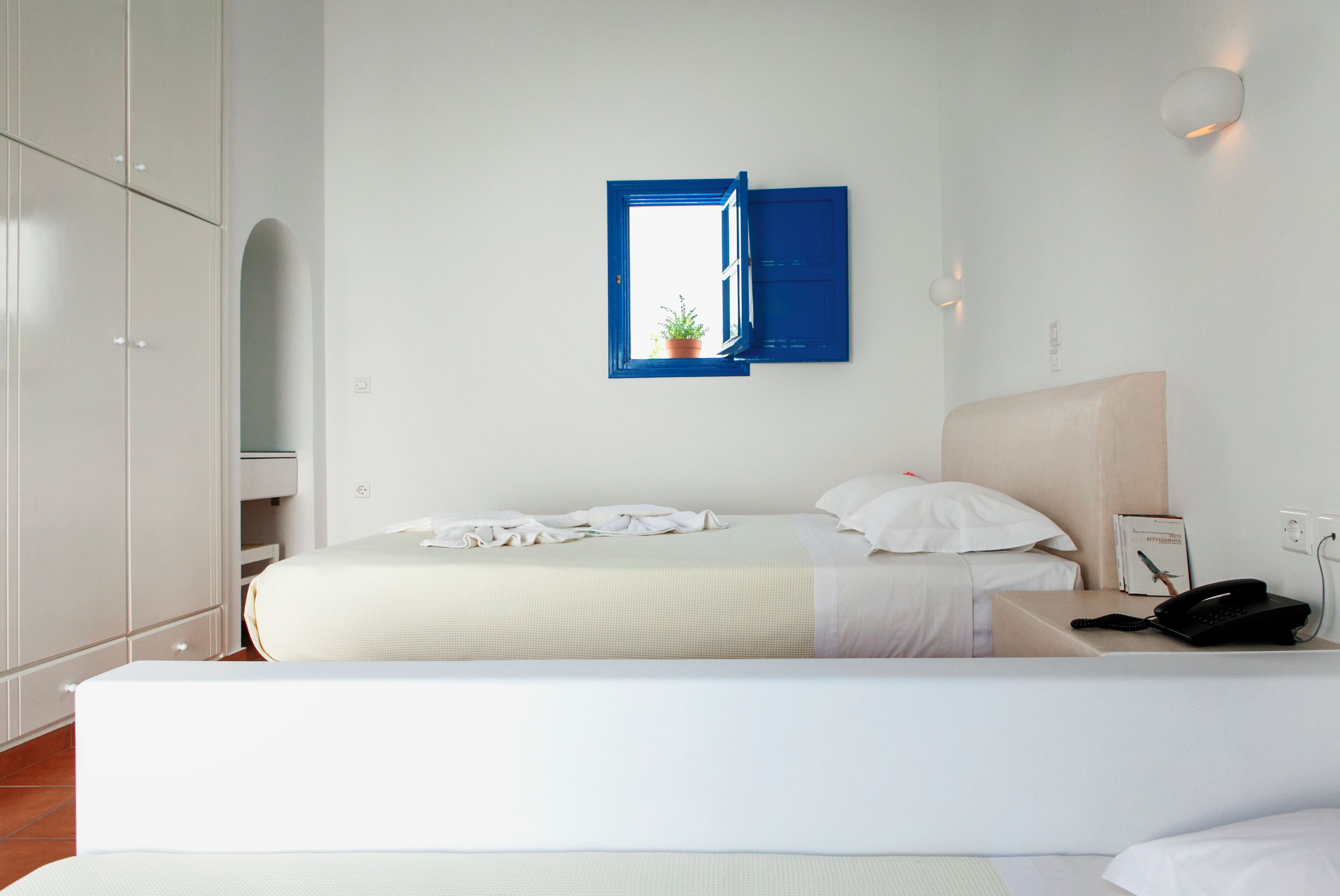 Habitación de hotel con dos camas, una ventana con contraventanas azules, una mesita de noche con una lámpara y un teléfono.