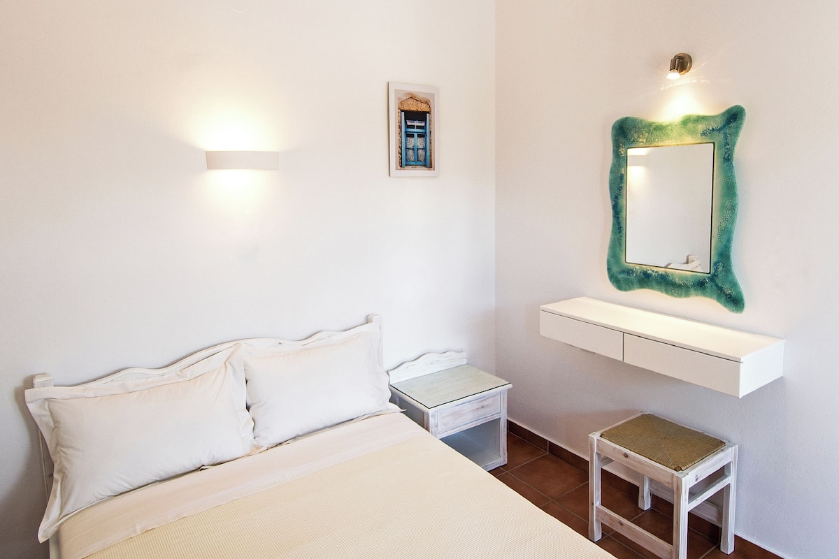A bedroom with a bed, bedside tables, a nightstand, a small stool, a wall-mounted mirror, and a shelf with a framed picture.