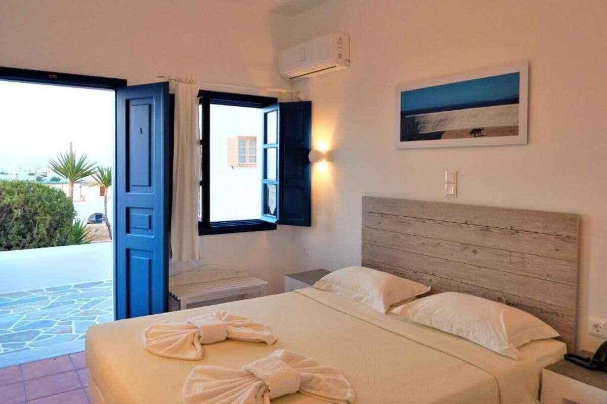A hotel room with a bed, a nightstand, an air conditioning unit, and a view of the outside.