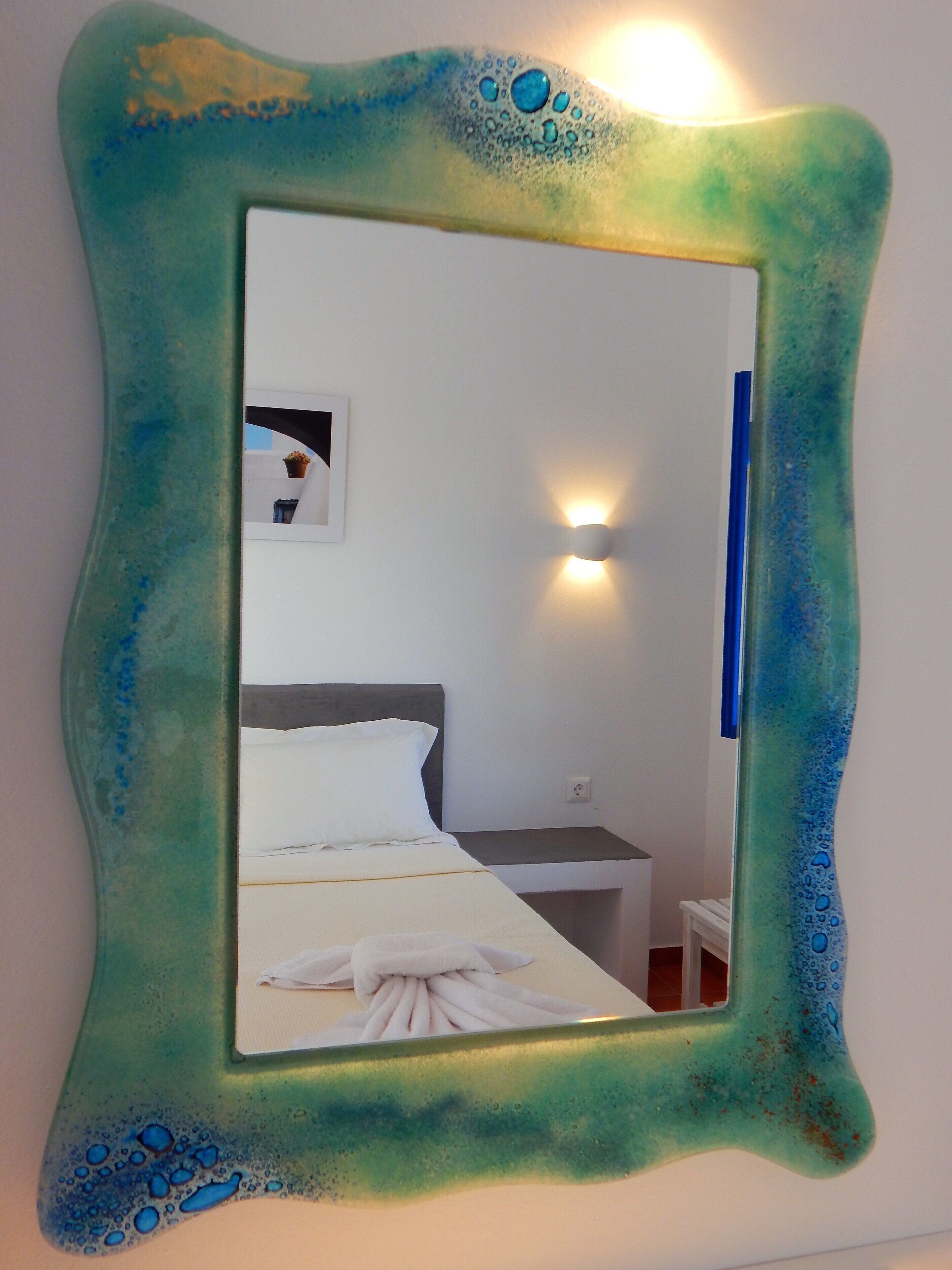 Un espejo con marco ondulado refleja una habitación con cama, mesita de noche y una lámpara fijada a la pared.