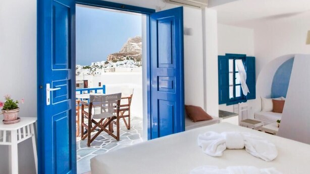 A hotel room with a bed, a table, and chairs. There is a view of a white building and a mountain through the open door. - Aegeo Hotel (Folegandros)