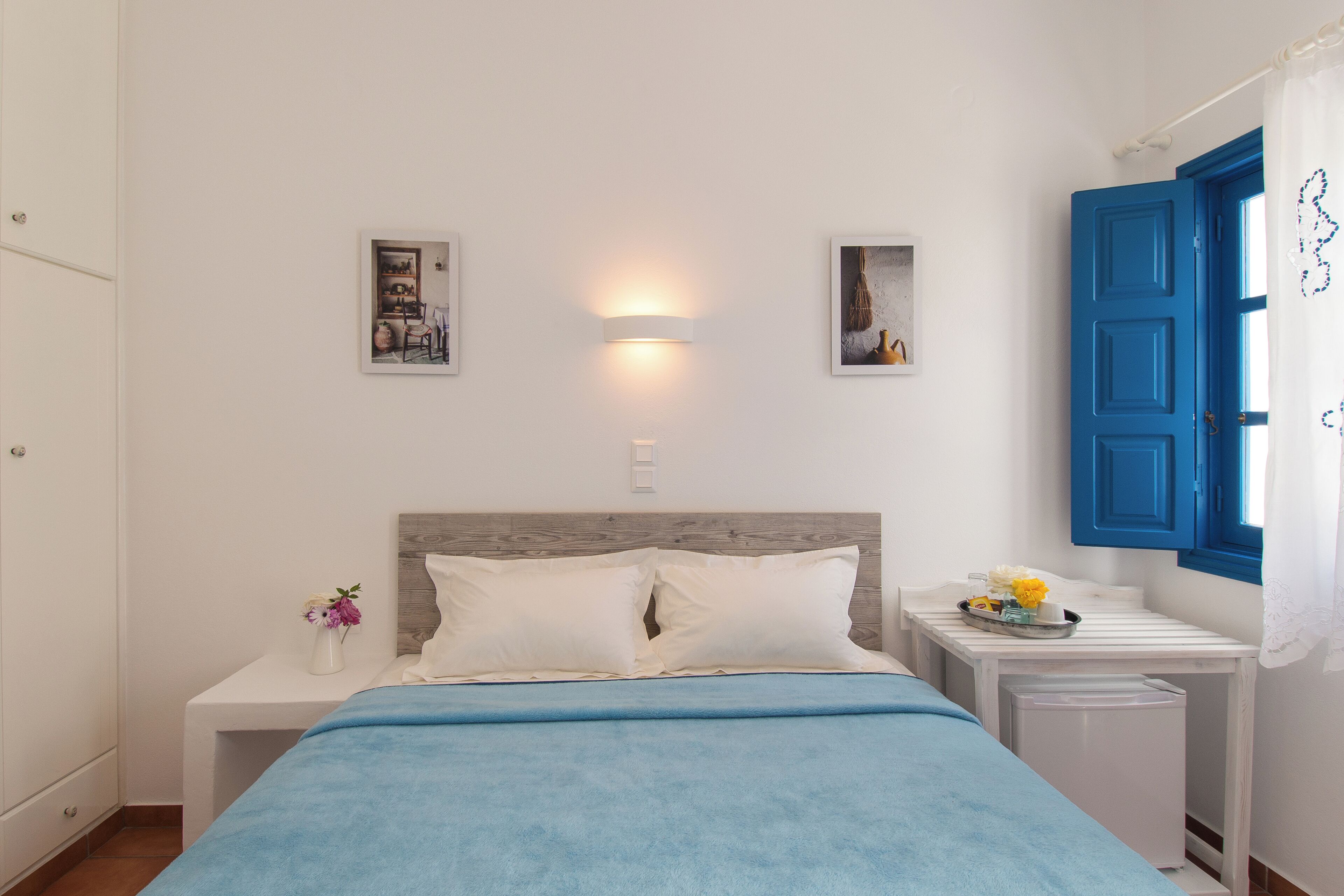 A bedroom with a bed, a nightstand with a vase of flowers, a wall-mounted light, and a window with blue shutters.