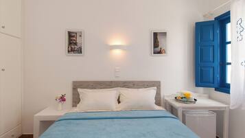 A bedroom with a bed, a nightstand with a vase of flowers, a wall-mounted light, and a window with blue shutters.