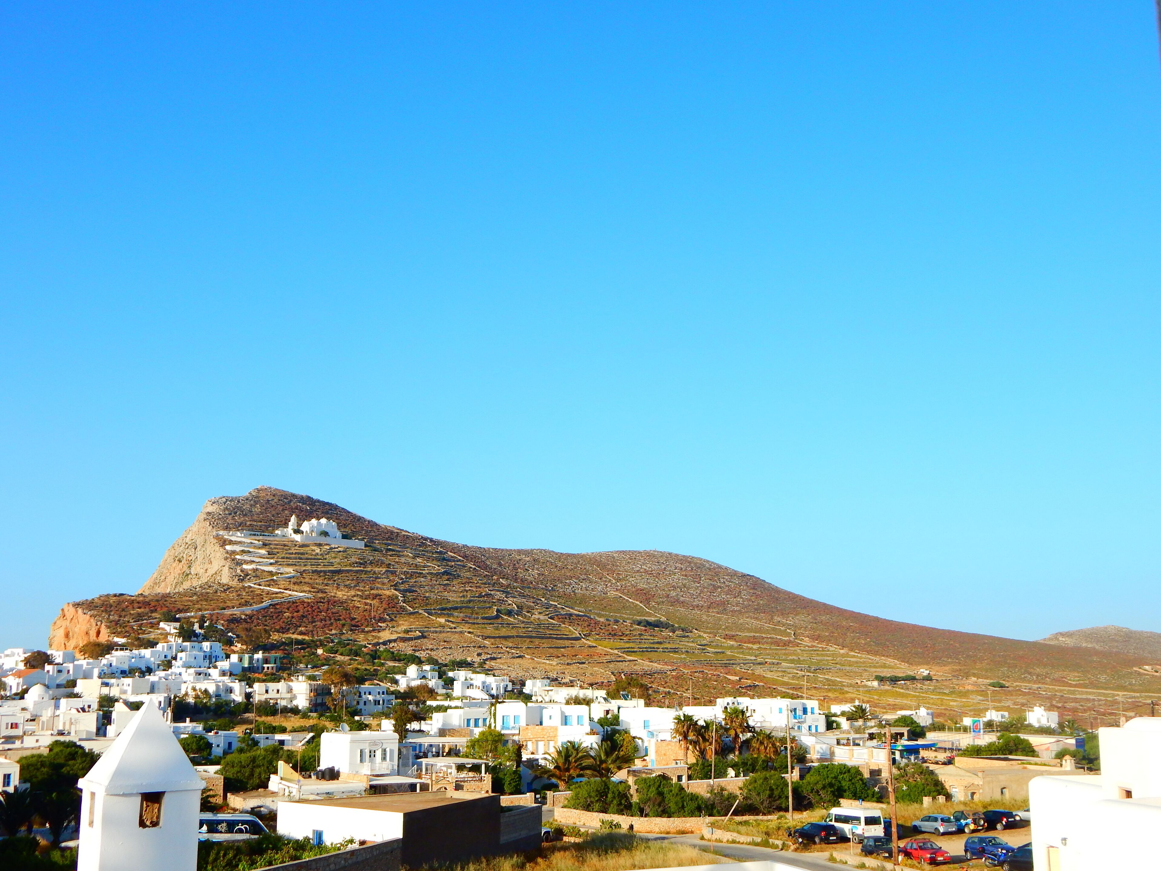 A coastal town with white buildings, a prominent hill, and a clear blue sky.