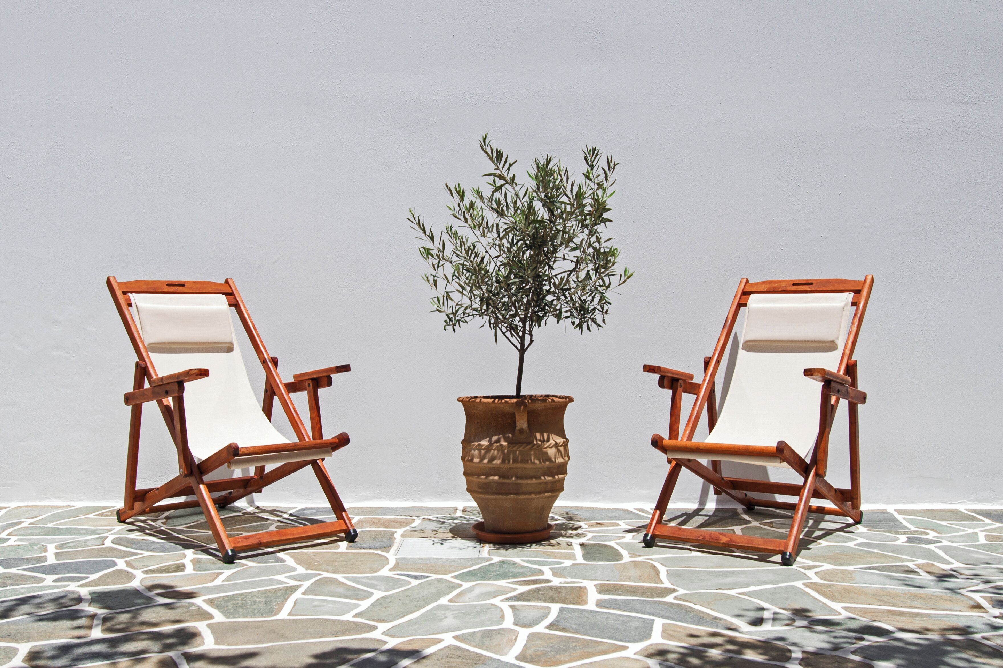 Zwei Holz-Loungesessel mit weißen Kissen, ein Blumentopf und ein Fliesenboden vor einer schlichten Wand.