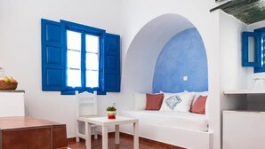 A room with a white sofa, a small table with a plant, a window with blue shutters, and a tiled floor. - Aegeo Hotel (Folegandros)