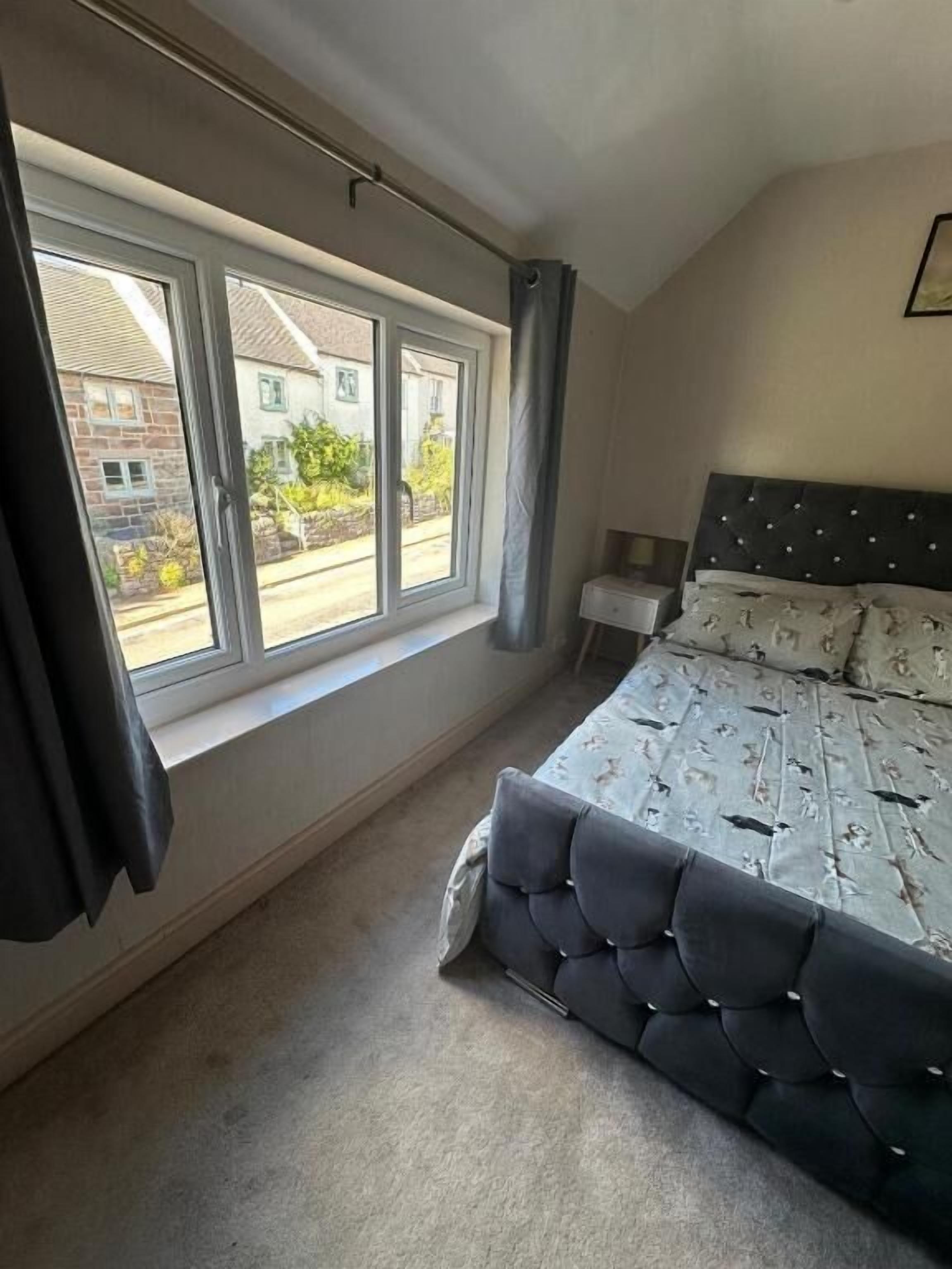 A bedroom with a large window, a bed with a tufted headboard, and a bedside table.