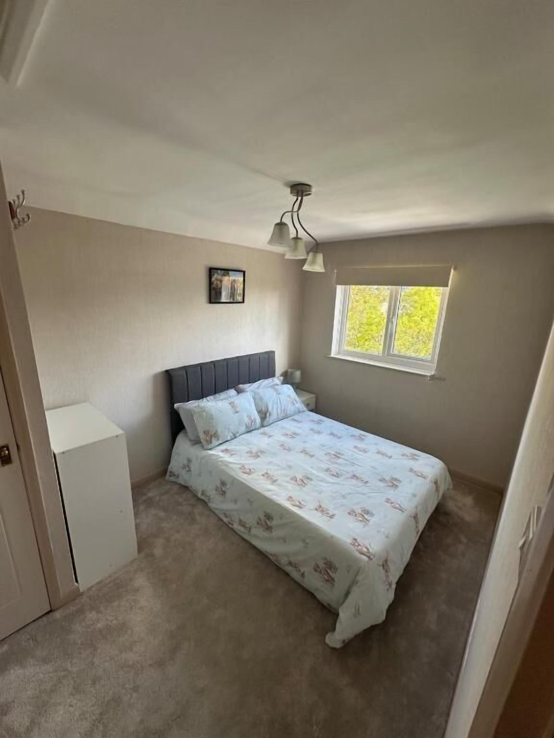 Een slaapkamer met een bed, nachtkastje, raam en een plafondlamp.