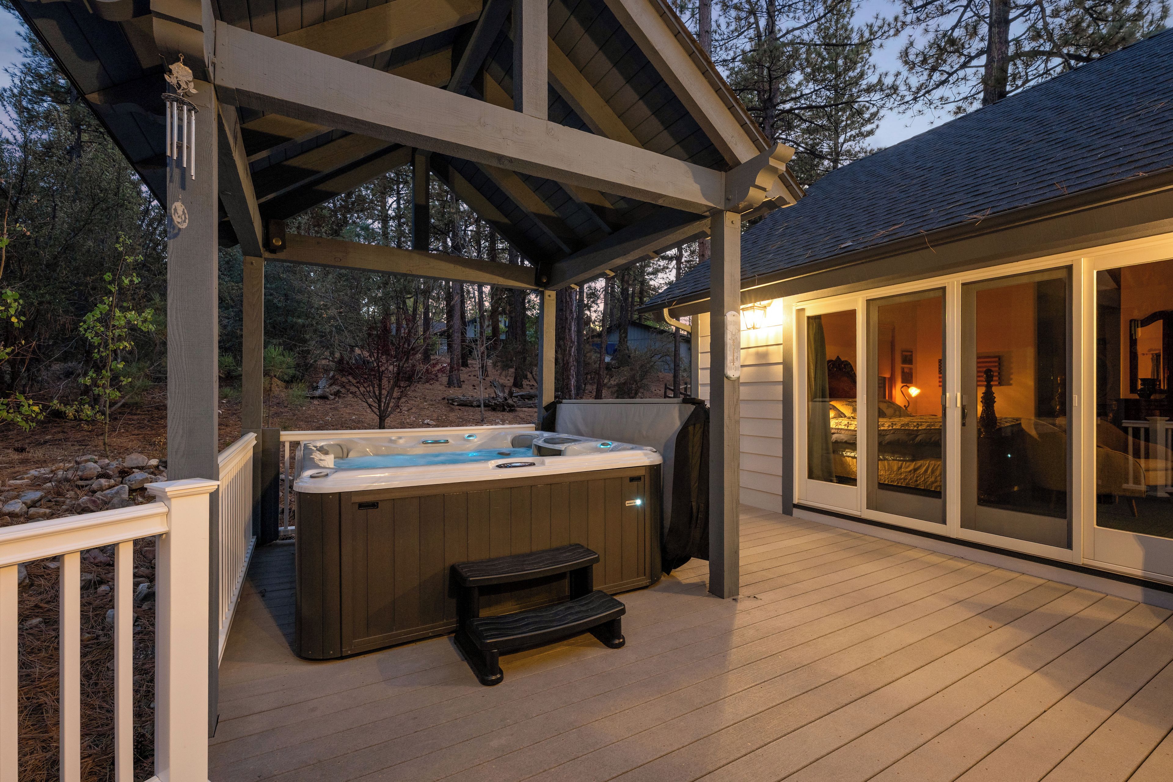 A hot tub under a wooden deck with a bench, surrounded by a forested area.