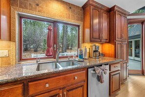 A kitchen with granite countertops, wooden cabinets, a double sink, and a window overlooking a wooded area. - Private Hot Tub, Game Room, 3,450 sq ft, Beautiful Home! Close to Everything (Big Bear City)