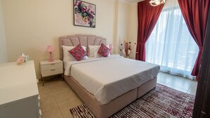 A bedroom with a bed, nightstands, a desk, a painting, and a window with curtains. - ina Homes - 1 BHK Tower 5 (Dubai)
