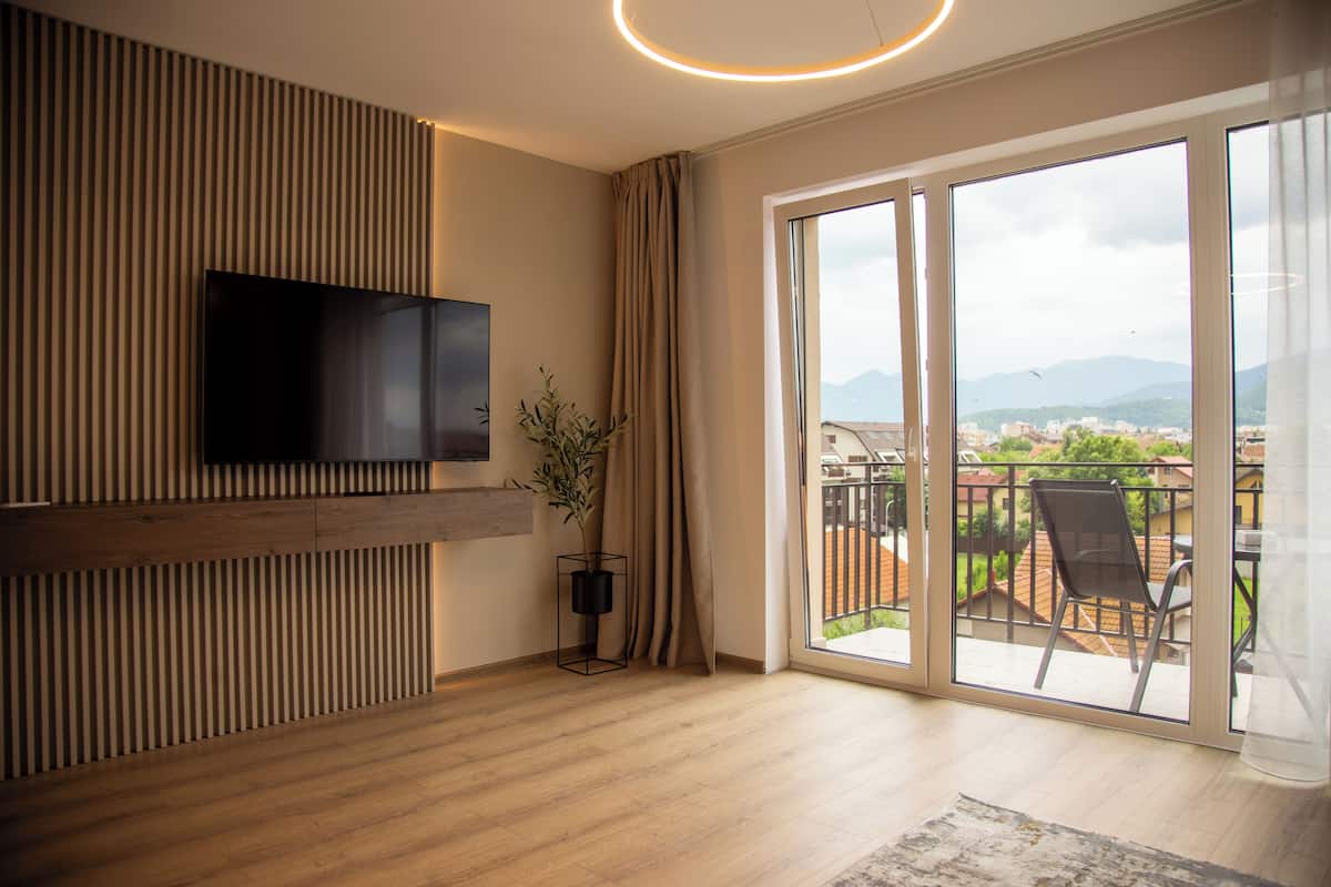 Un salon moderne doté d’un téléviseur à écran plat, d’une plante en pot et d’un balcon offrant une vue sur la ville et les montagnes.