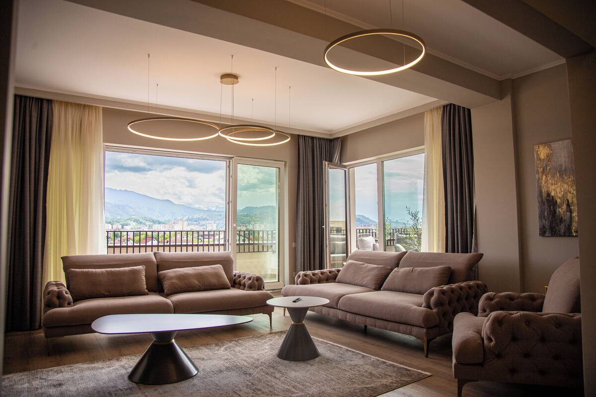Un vaste salon agrémenté d’un ensemble de meubles modernes, dont un canapé, une table basse et de grandes fenêtres offrant une vue sur la ville et les montagnes.