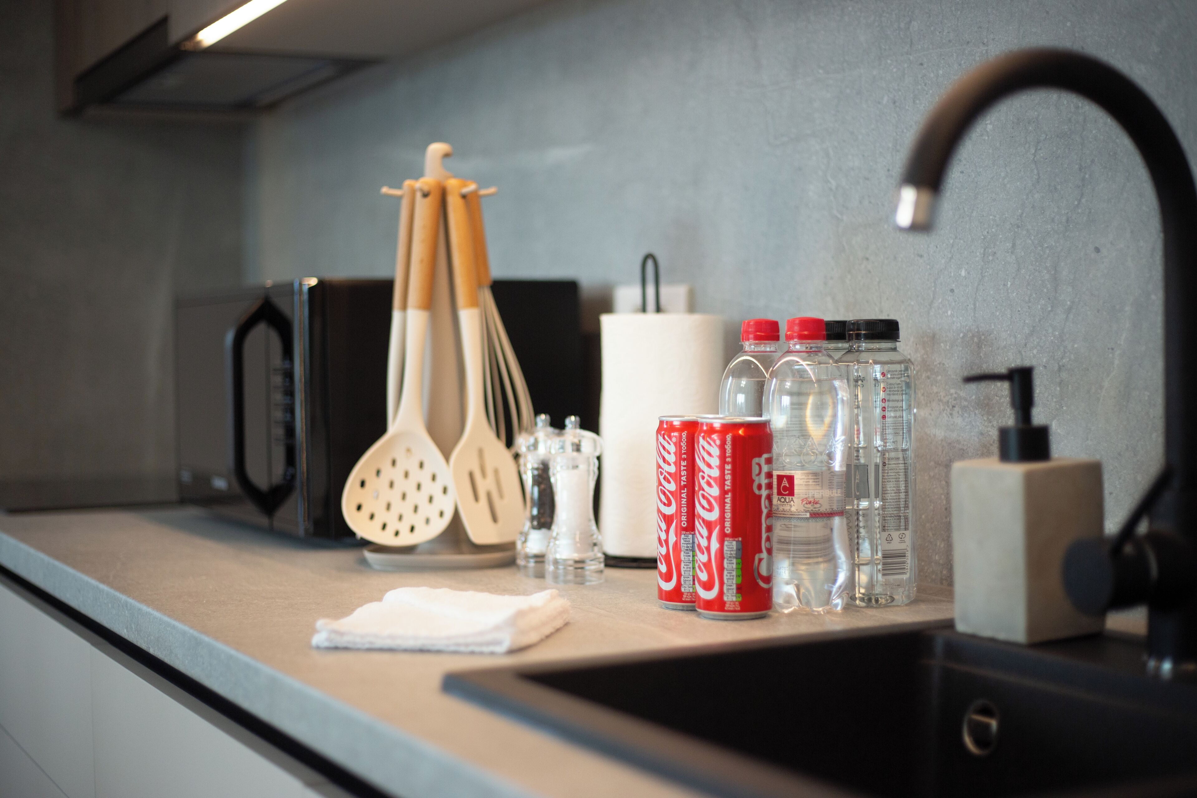 A kitchen counter with a microwave, wooden utensils, a white paper towel holder, and two Coca-Cola cans.