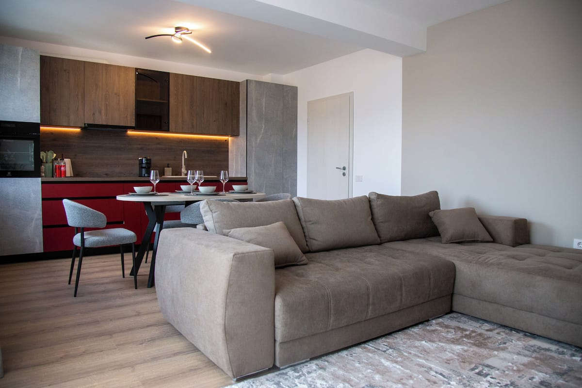 Un salon moderne avec un canapé d’angle, un coin repas comprenant une table et des chaises, et une cuisine dotée de meubles en bois et d’un plan de travail rouge.