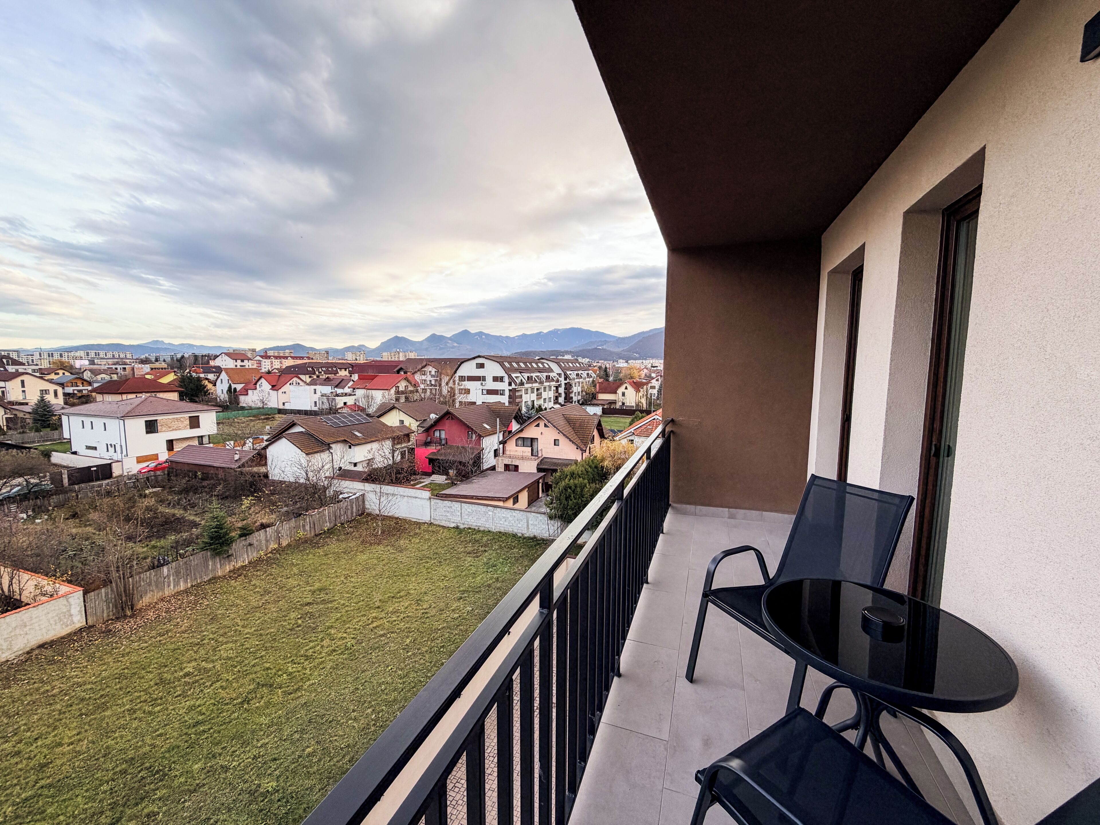 A balcony with a table and chairs, overlooking a residential area with houses and mountains in the distance.