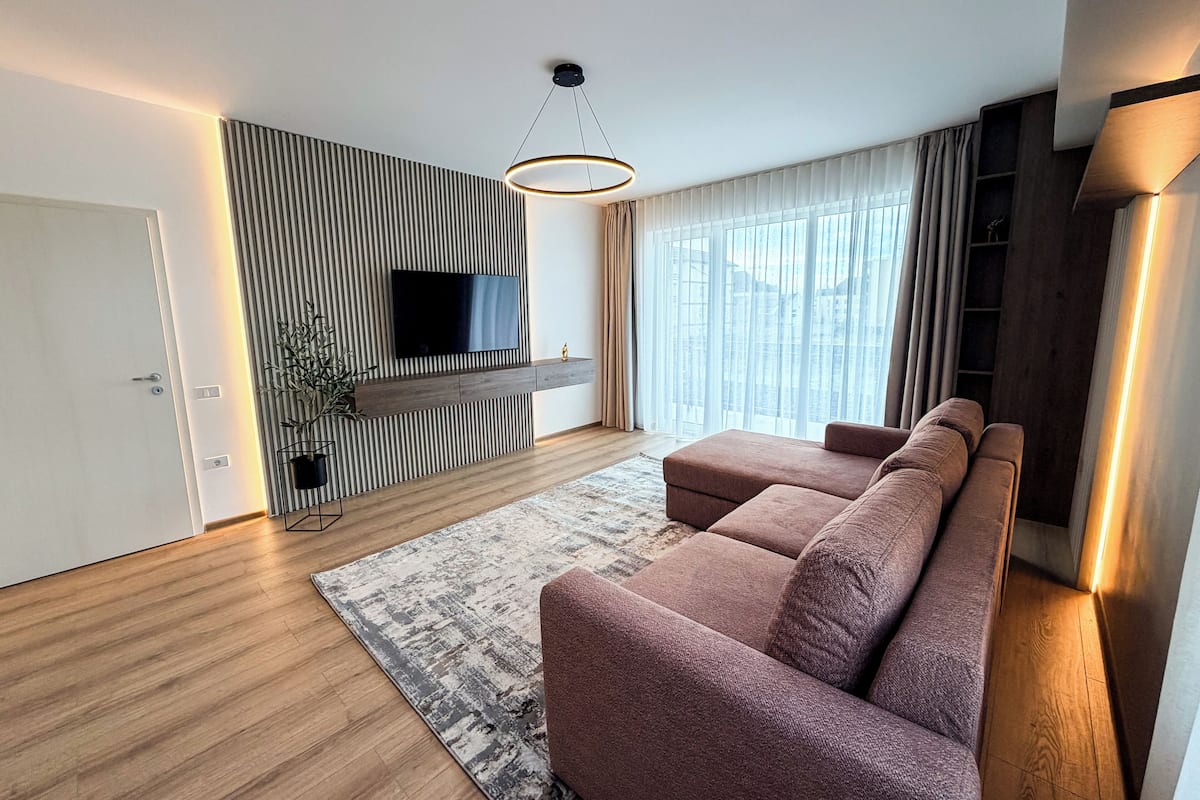 Un salon moderne doté d’un téléviseur à écran plat, d’un canapé, d’un plancher en bois et d’une grande fenêtre avec des rideaux.