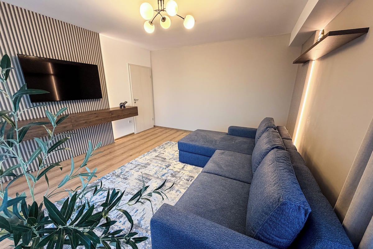 Un salon moderne avec un canapé d’angle, un téléviseur à écran plat, un mur décoratif en bois et un luminaire au plafond.